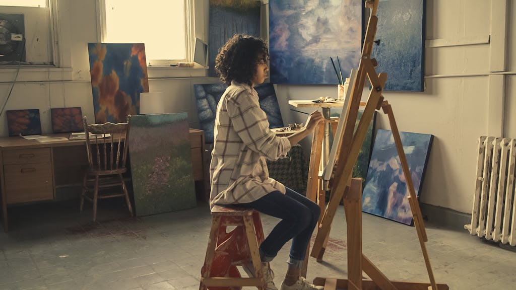 A young woman focused on painting in an art studio filled with her work.